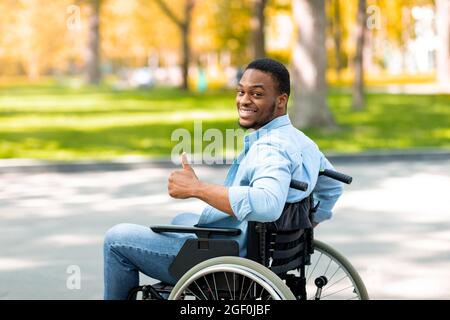 Heureux handicapé noir en fauteuil roulant passer du temps au parc de la ville en automne, montrant le pouce vers le haut et souriant Banque D'Images