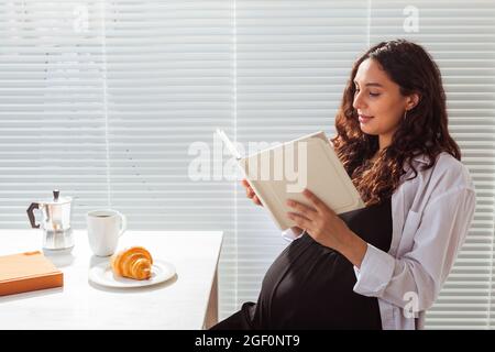 Femme enceinte mangeant et lisant le livre à la maison. Concept matin, petit déjeuner et grossesse Banque D'Images