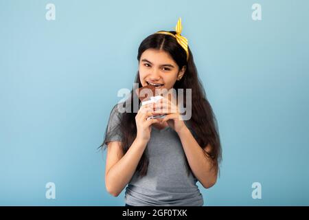 Adorable adolescente indienne mangeant une savoureuse barre de chocolat, en succombant à la tentation sur fond de studio bleu Banque D'Images