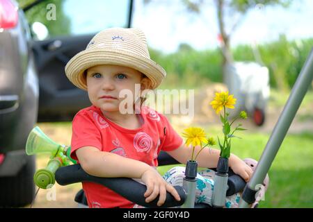 Gros plan d'une petite fille mignonne dans un chapeau. Portrait d'une jolie petite fille dans un chapeau d'été regardant l'appareil photo assis sur un vélo. Banque D'Images