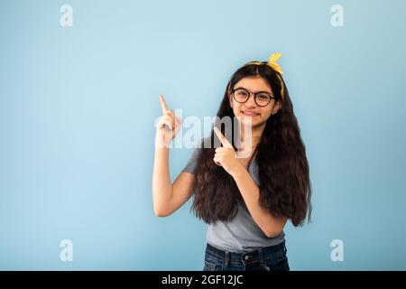 Offre sympa. Une adolescente indienne gaie pointant deux doigts de côté à l'espace de copie sur fond bleu de studio Banque D'Images