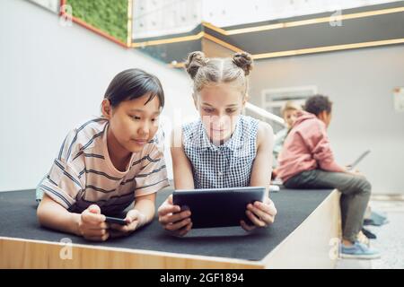 Portrait de deux adolescentes utilisant des gadgets tout en se relaxant dans l'intérieur moderne de l'école, l'espace de copie Banque D'Images