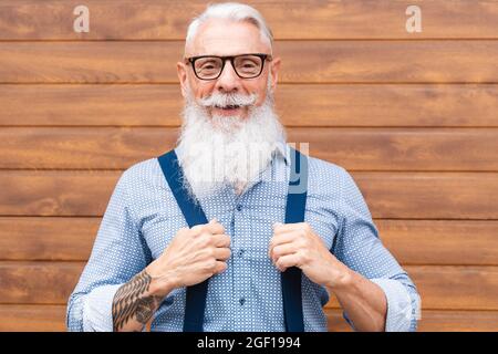 Joyeux hipster homme senior souriant sur la caméra avec mur de bois en arrière-plan à l'extérieur dans la ville - Focus sur le visage Banque D'Images