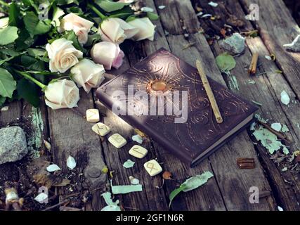 Encore la vie avec des runes, livre de sorcière et roses blanches sur des planches dans le jardin. Arrière-plan ésotérique, gothique et occulte, concept mystique et wicca d'Halloween Banque D'Images