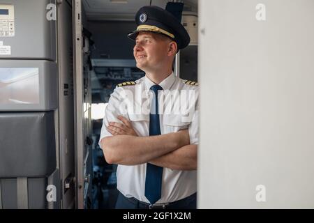 Un pilote masculin rêveur en uniforme et en chapeau tenant les bras croisés et souriant loin en posant, debout à l'intérieur de l'avion Banque D'Images