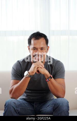 Portrait d'un jeune homme heureux assis sur un canapé à la maison, souriant et regardant l'appareil photo Banque D'Images