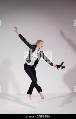 Une ballerine pieds nus gracieuse dans un costume d'affaires sautant avec des chaussures dans ses mains sur un fond blanc. Banque D'Images