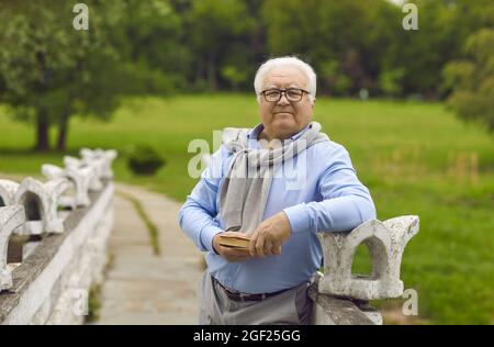 Un homme âgé se tient dans le parc avec un livre entre ses mains. Banque D'Images