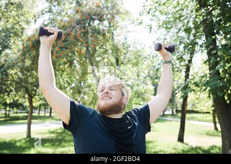 Homme en surpoids dans les vêtements de sport faisant des exercices avec des haltères à l'extérieur Banque D'Images