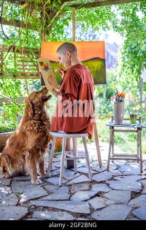 Jeune artiste femme travaillant sur son art toile peinture à l'extérieur dans son jardin avec Golden retriever gardant son entreprise. Concept de loisirs créatifs. Banque D'Images