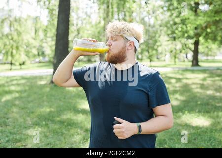 Un homme barbu en surpoids boit du jus frais à la bouteille après un entraînement sportif en plein air Banque D'Images