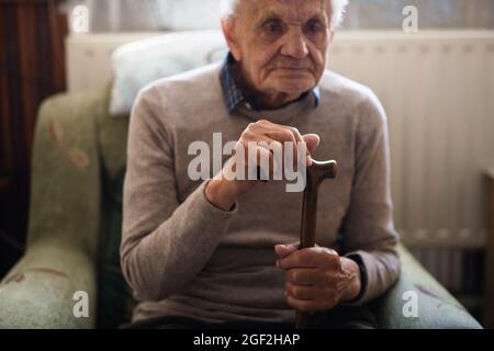 Triste homme âgé avec un bâton de marche assis sur un fauteuil à l'intérieur à la maison, se reposant. Banque D'Images