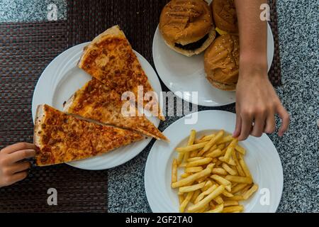 les mains des enfants pour prendre des repas rapides sur la table. Pizza, hamburger, frites. Banque D'Images