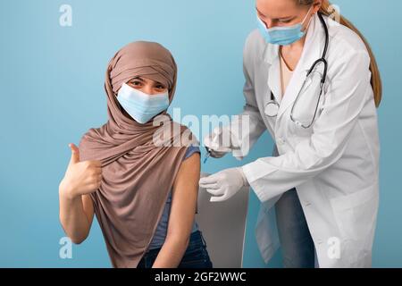 Jeune fille musulmane dans le hijab et masque médical se faire vacciner contre le covid par injection de la femme médecin, en faisant des gestes vers le haut Banque D'Images