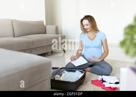 Femme enceinte en t-shirt bleu, liste de contrôle, téléphone parlant et préparation de la maternité à l'hôpital à la maison. Bonne grossesse emballage des vêtements de bébé dans une valise. Banque D'Images