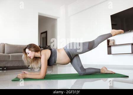 Sportive belle blonde femme enceinte s'exerçant sur un tapis de yoga vert dans des vêtements de sport gris à la maison. Une maternité sportive et saine. Fitness, maternité heureuse, concept de style de vie. Banque D'Images