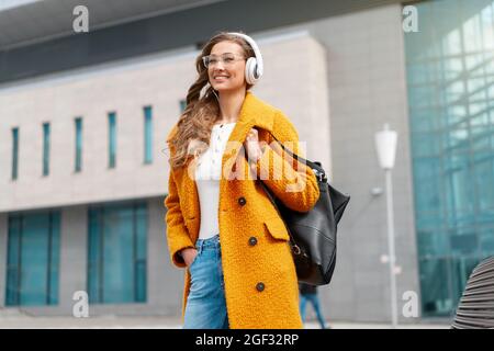 Femme porte des lunettes Ecouter musique casque marche en plein air City Street habillé élégant manteau jaune et sac à dos noir Smile Caucasien femelle 30s Enj Banque D'Images