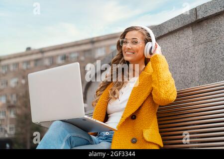 Femme d'affaires porter des lunettes Ecouter musique casque extérieur assis banc utilisant ordinateur portable extérieur habillé élégant manteau jaune sourire femelle caucasien 30 Banque D'Images