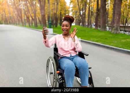 Bonne jeune femme noire handicapée en fauteuil roulant avec appel vidéo sur smartphone, agitant à la caméra dans le parc d'automne Banque D'Images
