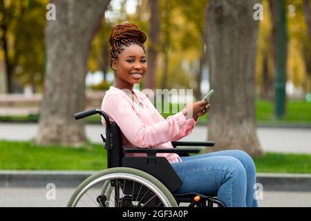 Heureuse femme noire handicapée en fauteuil roulant utilisant le téléphone portable, la vérification des médias sociaux, la navigation sur le Web à l'automne parc Banque D'Images