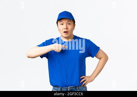 Surpris et choqué de coursier asiatique, livreur dans la casquette bleue uniforme et t-shirt, regardant confus et pointant sur lui-même inquiet, étant nommé Banque D'Images