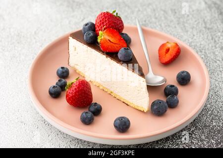 Un morceau de cheesecake recouvert de chocolat avec des bleuets et des fraises sur une assiette rose avec une cuillère à dessert Banque D'Images