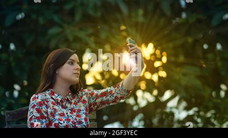 Belle jeune femme latine en robe à motif floral prenant un selfie avec son smartphone dans le jardin au coucher du soleil avec les rayons du soleil passant par le Banque D'Images