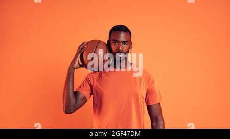 Un joueur de basket-ball sérieux qui regarde l'appareil photo. Portrait d'un homme avec une balle de panier sur son épaule contre un fond orange. Banque D'Images