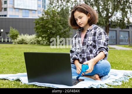 Une jeune fille adorable assise sur l'herbe et travaillant sur un ordinateur portable. Une écolière regarde une classe Internet et étudie à distance. La fille utilise le sans fil Banque D'Images