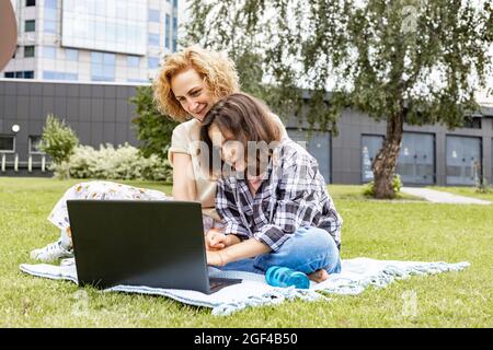 Une jeune fille adorable est assise sur l'herbe dans le parc avec sa mère et utilise un ordinateur portable. Une écolière avec sa mère regarde Internet et étudie rem Banque D'Images