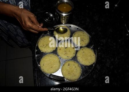 Femme cuisine à la main faisant populaire sud indien plat à la vapeur rava idli ou semolina idli. Verser le ghee ou le beurre clariné sur la préparation de la cuisine de l'idlis Banque D'Images