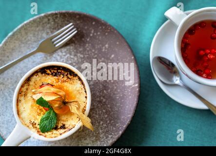 Délicieux dessert crème brûlée décoré de baies fraîches et de feuilles de menthe et de tisane aux canneberges sur fond gris et vert. Style de pose à plat. Maquette pour café ou restaurant avec espace de copie. Banque D'Images