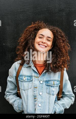 Jeune femme souriante aux cheveux bouclés debout avec les mains dans les poches Banque D'Images