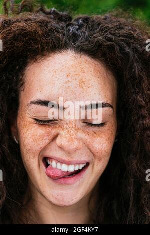 Jeune femme gaie aux cheveux bouclés avec les yeux fermés qui collent hors de la langue Banque D'Images