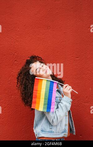 Jeune femme souriante debout avec un drapeau arc-en-ciel devant le mur rouge Banque D'Images