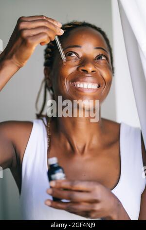 Femme afro souriante regardant tout en appliquant du sérum de visage Banque D'Images