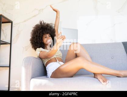 Femme afro avec la main levée tenant une tablette numérique tout en étant assise sur un canapé à la maison Banque D'Images
