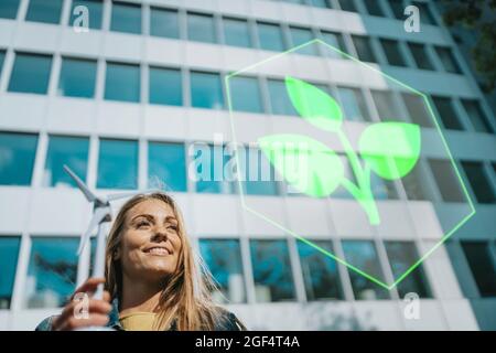 Femme souriante avec le modèle de moulin à vent regardant le logo de plante près du bâtiment Banque D'Images