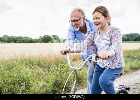 Homme mûr souriant aidant une fille à faire du vélo dans la prairie Banque D'Images