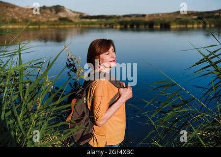 Femme avec les yeux fermés tenant sac à dos tout en se tenant près du lac Banque D'Images