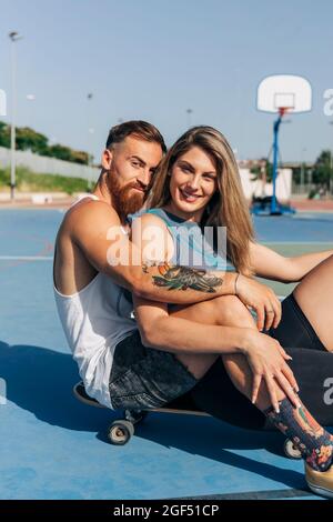 Jeune femme souriante assise avec un petit ami sur un skateboard au terrain de basket-ball Banque D'Images