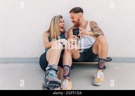 Homme joyeux tenant un smartphone tout en riant avec une petite amie penchée sur le mur Banque D'Images