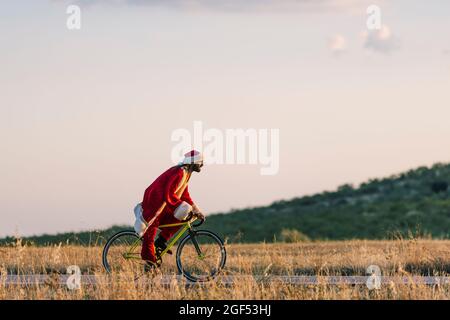 Jeune homme en costume du Père Noël à vélo sur la route au coucher du soleil Banque D'Images