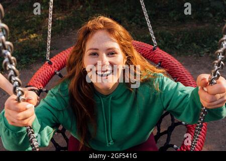 Bonne jeune femme à tête rouge appréciant son swing sur l'aire de jeux Banque D'Images