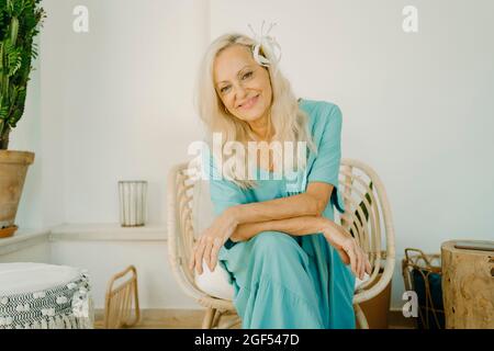 Femme âgée avec fleur dans de longs cheveux blancs assis sur une chaise à la maison Banque D'Images
