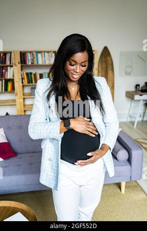 Bonne jeune femme enceinte freelancer touchant l'abdomen tout en étant debout à la maison Banque D'Images