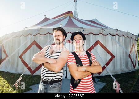 Un artiste de construction musclé debout avec les bras croisés par le clown devant la tente de cirque Banque D'Images
