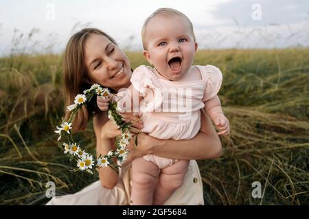 Mère souriante tenant bébé fille joyeuse avec tiara de fleur dans le champ Banque D'Images