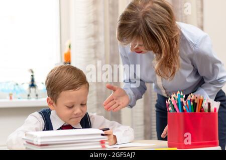 Un jeune garçon de première classe mignon dans un uniforme d'école à la maison joue sur un smartphone à un bureau avec des livres et des crayons. Maman le grimpa. Mise au point sélective. Gros plan. Banque D'Images