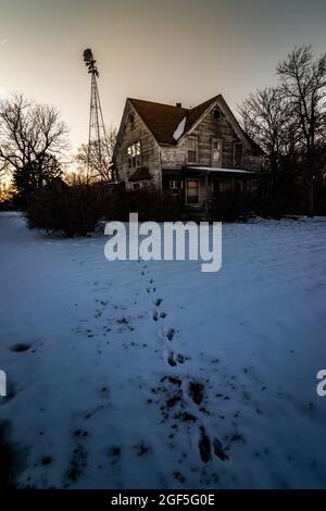 Une photo verticale d'une maison abandonnée et d'un vieux moulin à vent au coucher du soleil, avec des empreintes de pas dans la neige menant à la maison en hiver Banque D'Images